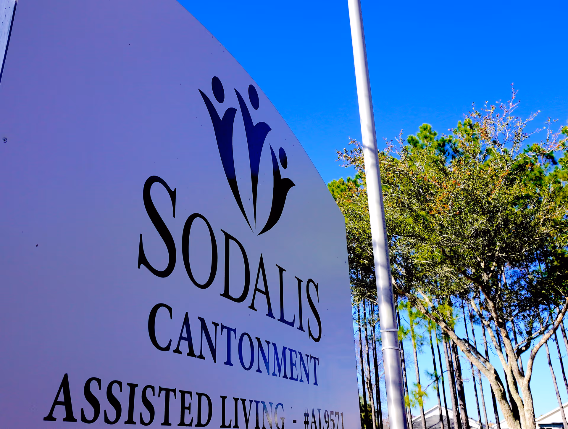 A large white sign for Sodalis Cantonment Assisted Living with a logo of three stylized human figures, set against a clear blue sky and trees in the background.