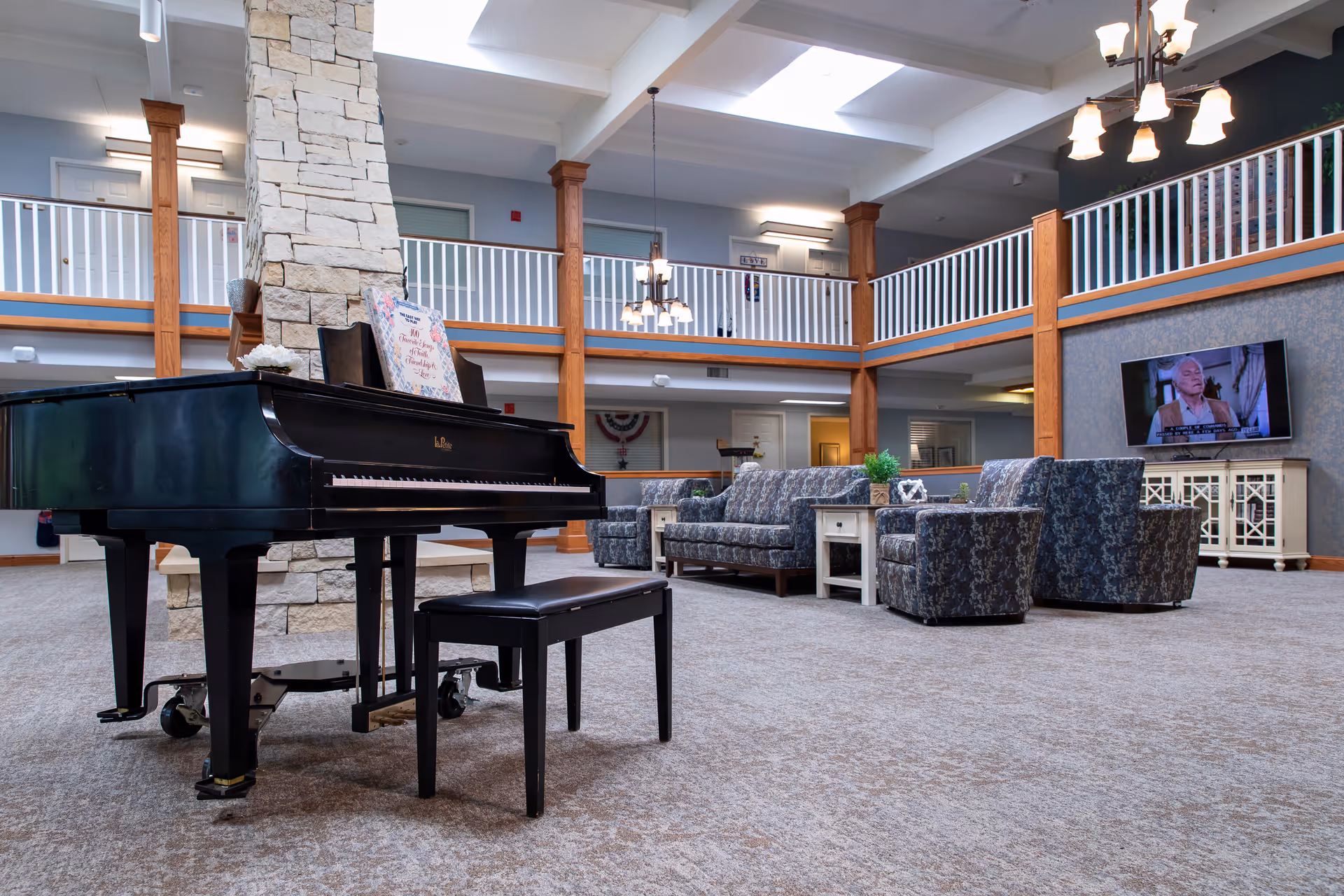 Spacious senior living facility common area with a black grand piano and matching bench in the foreground. Behind the piano is a stone fireplace extending to the ceiling. The room features a seating area with patterned armchairs and a sofa around a white coffee table. A flat-screen TV is mounted on the wall above a white cabinet. The room has high ceilings with exposed beams, wooden pillars, and a second-floor balcony with white railings. Multiple light fixtures hang from the ceiling providing warm lighting.