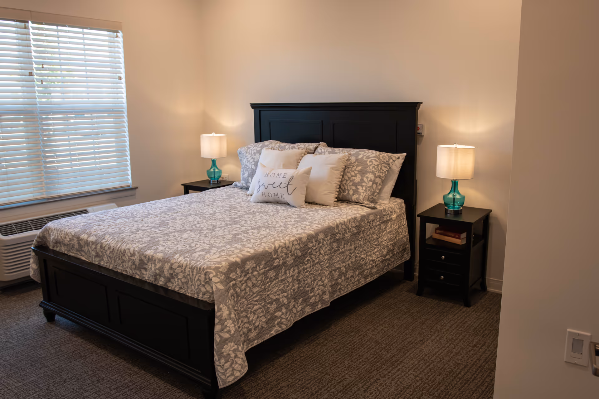Dark wood bed with patterned gray comforter, two nightstands, lamps, and a window with blinds.