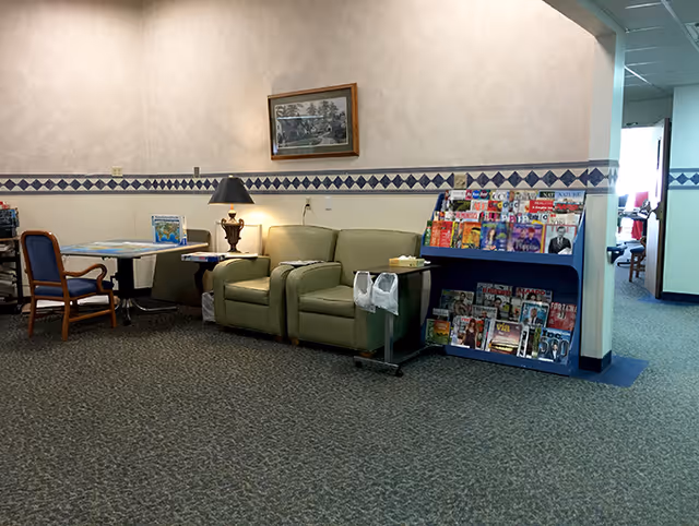 Interior common area with a green loveseat, a side table with a lamp, a magazine rack filled with various magazines, a table with chairs, and a framed picture on the wall. The space has carpeted flooring and wallpaper with a diamond pattern border.