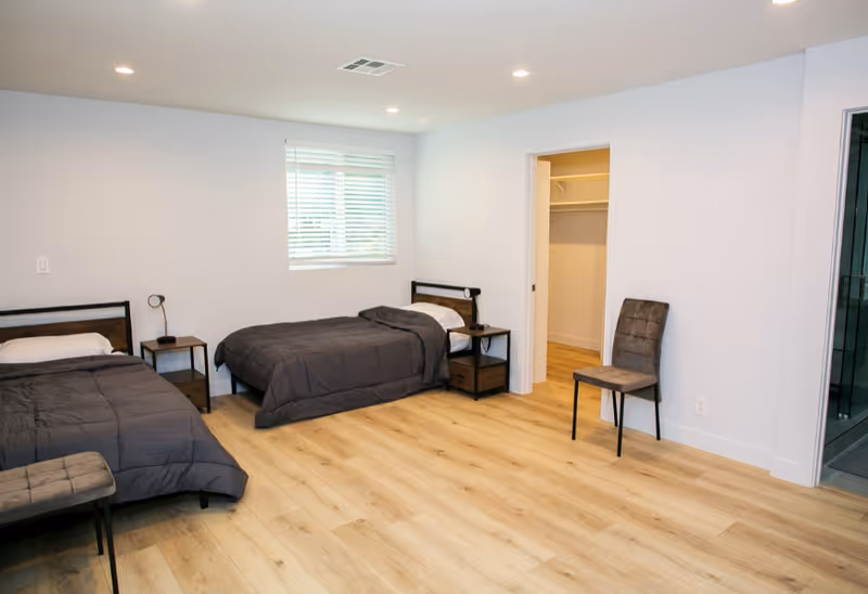 Two twin beds with dark gray comforters, nightstands, and an open closet in a bright bedroom.