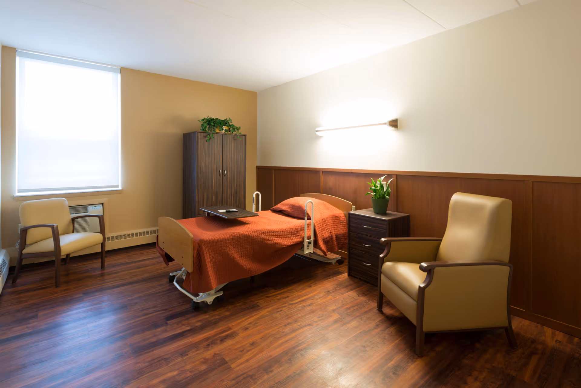 A furnished senior living bedroom with a bed, chair, nightstand, and orange bedspread.