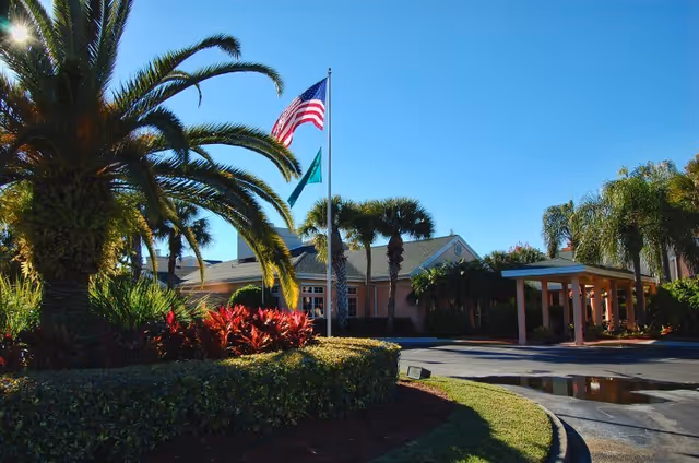 Exterior view of The Gardens of Port St. Lucie entrance with flags and palm trees.