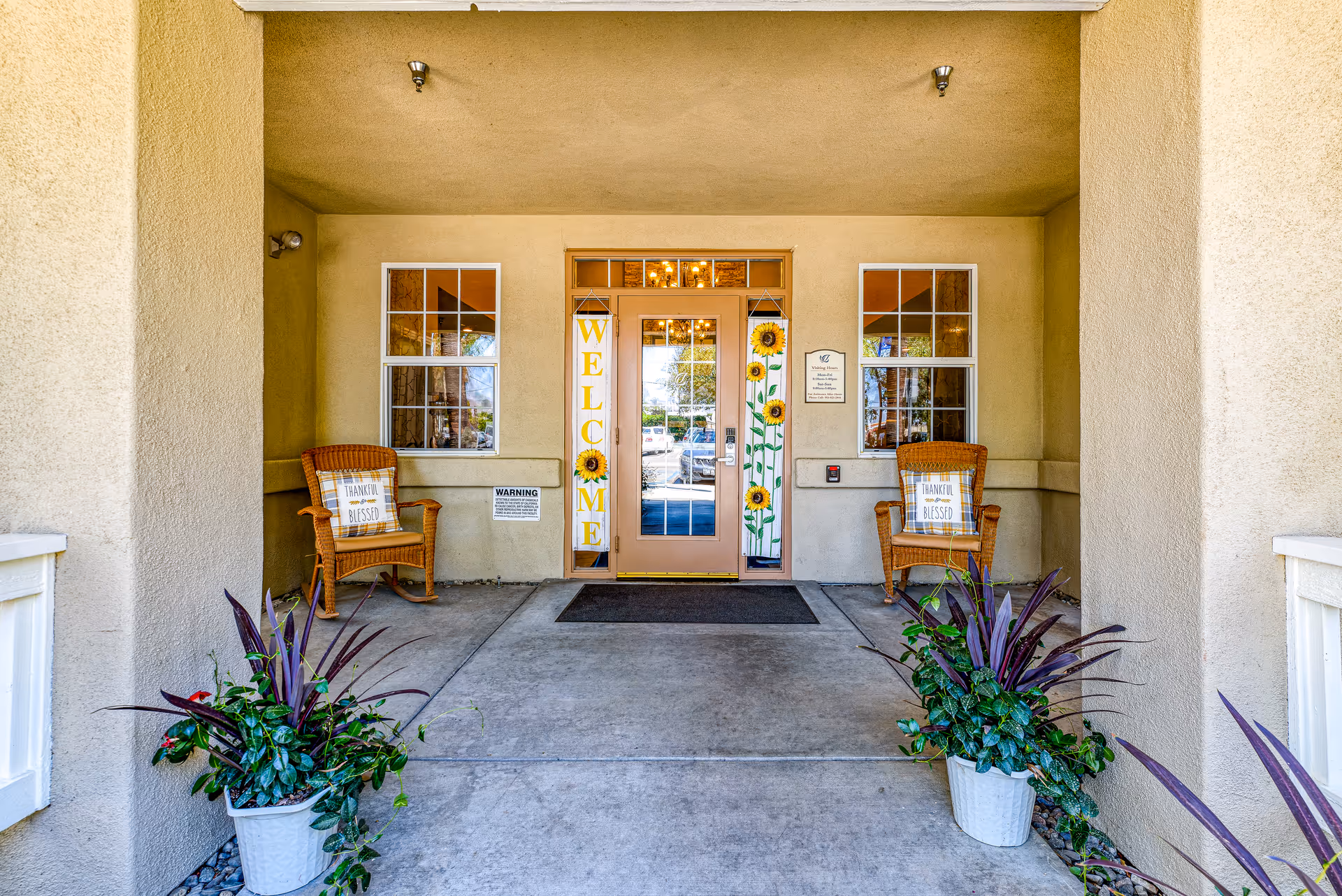 Covered entrance area of a building with a glass door decorated with sunflower-themed welcome banners. Two wicker chairs with cushions that say 'Thankful & Blessed' are placed on either side of the door. Two potted plants with green and purple leaves are positioned near the entrance. The walls and ceiling are beige, and there are two windows flanking the door.