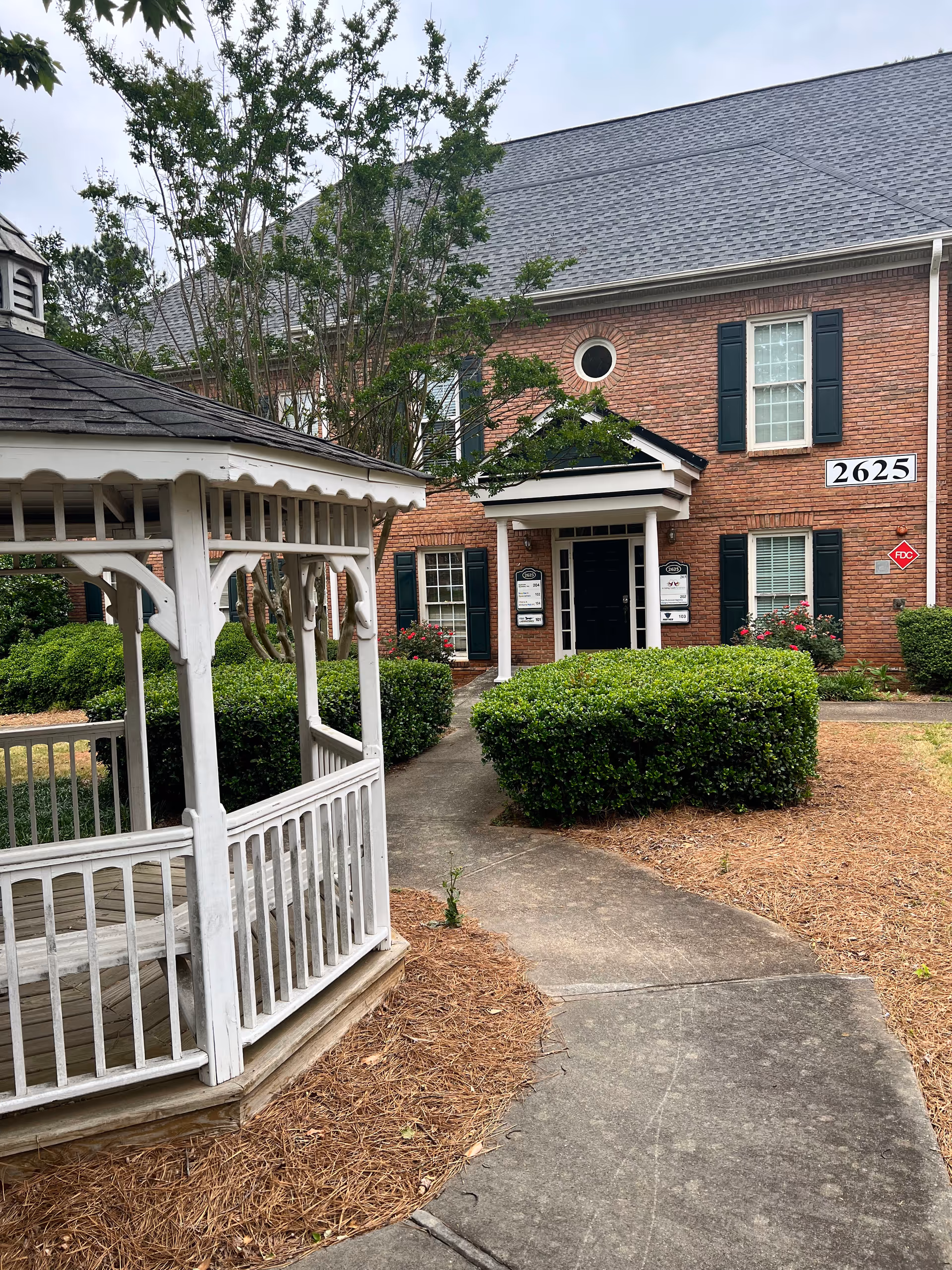 Brick senior living building with white gazebo, walkway, and manicured hedges outdoors.