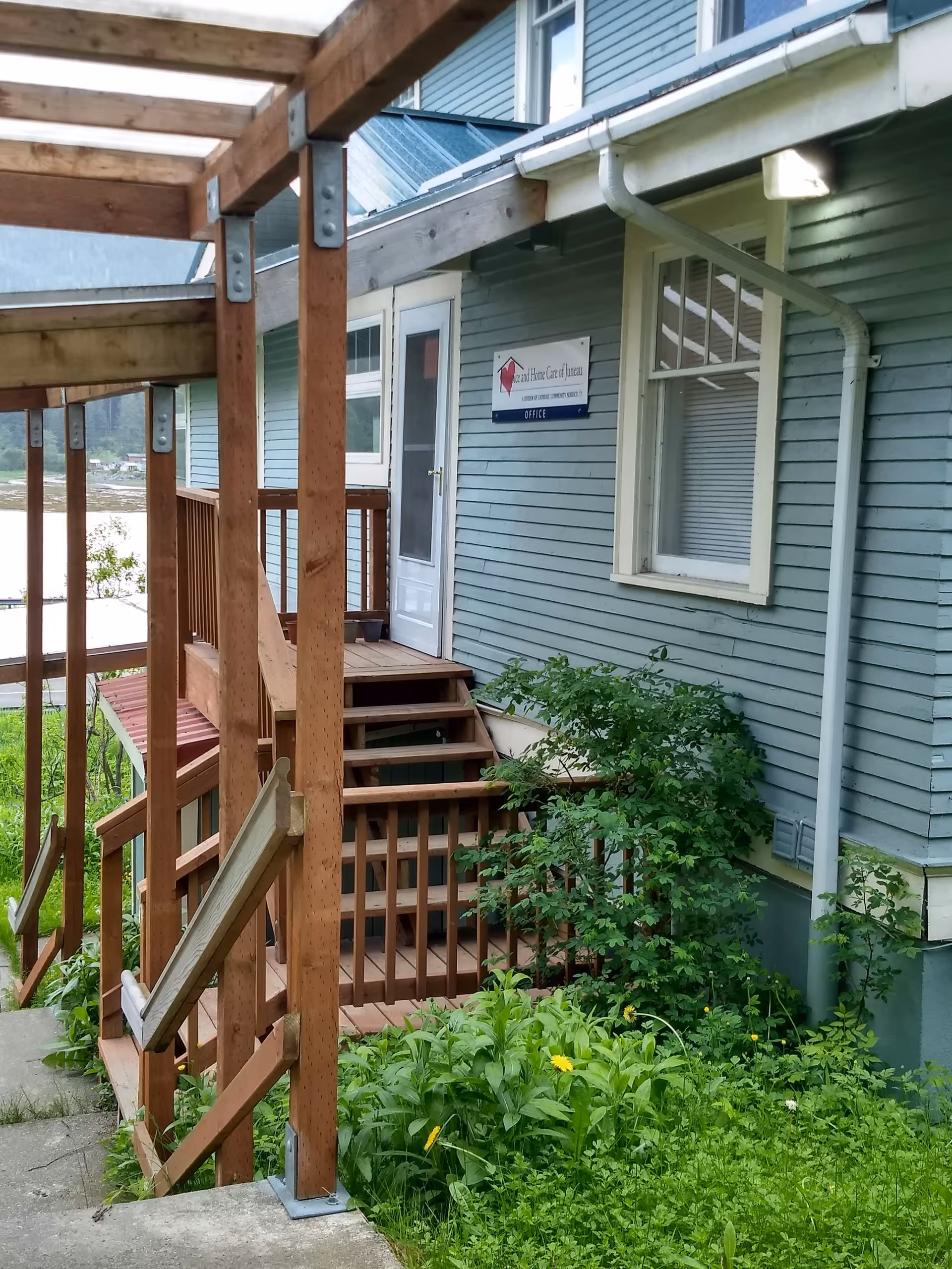 Exterior of a blue Hospice building with wooden stairs and greenery.