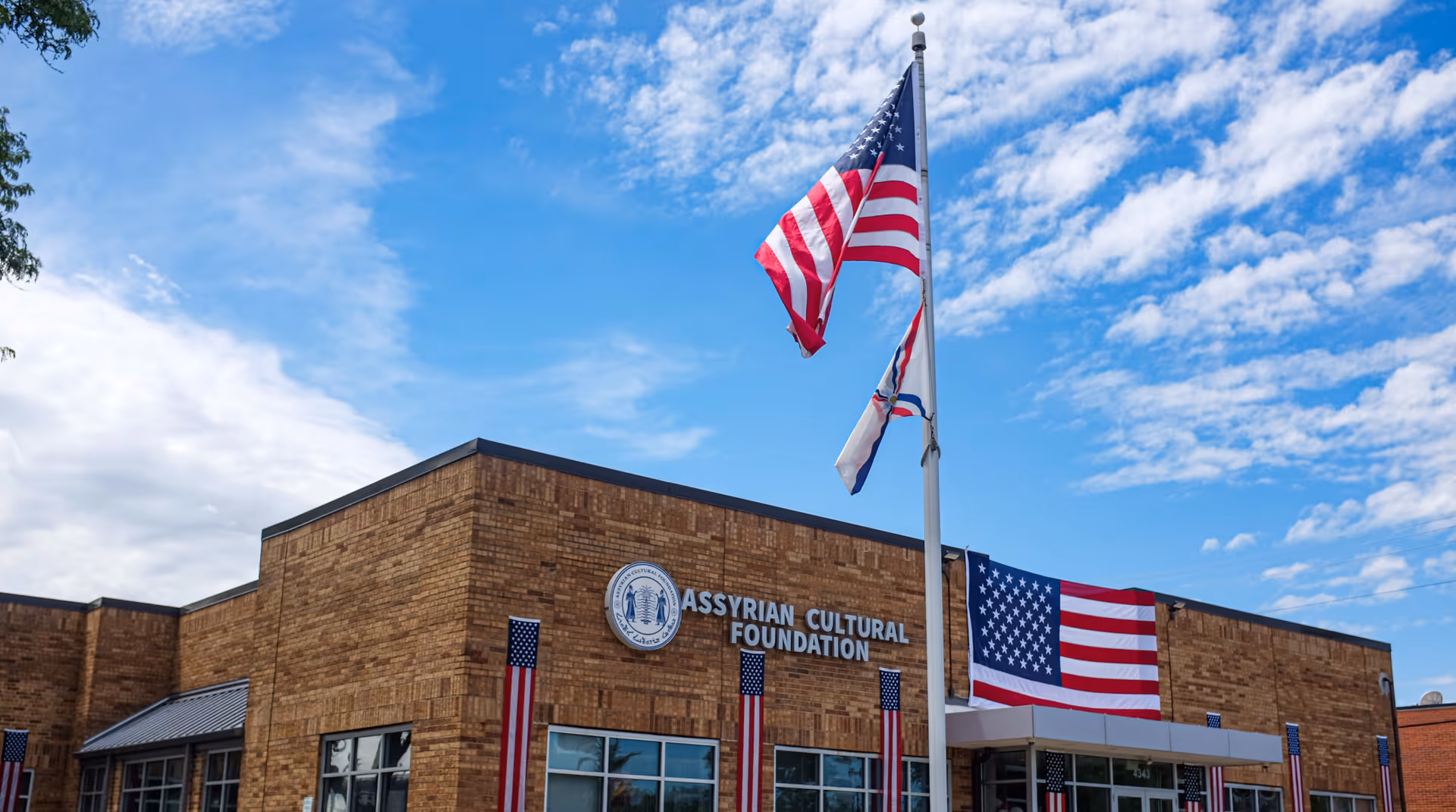 Brick facility exterior with Assyrian Cultural Foundation sign and American flags against blue sky.