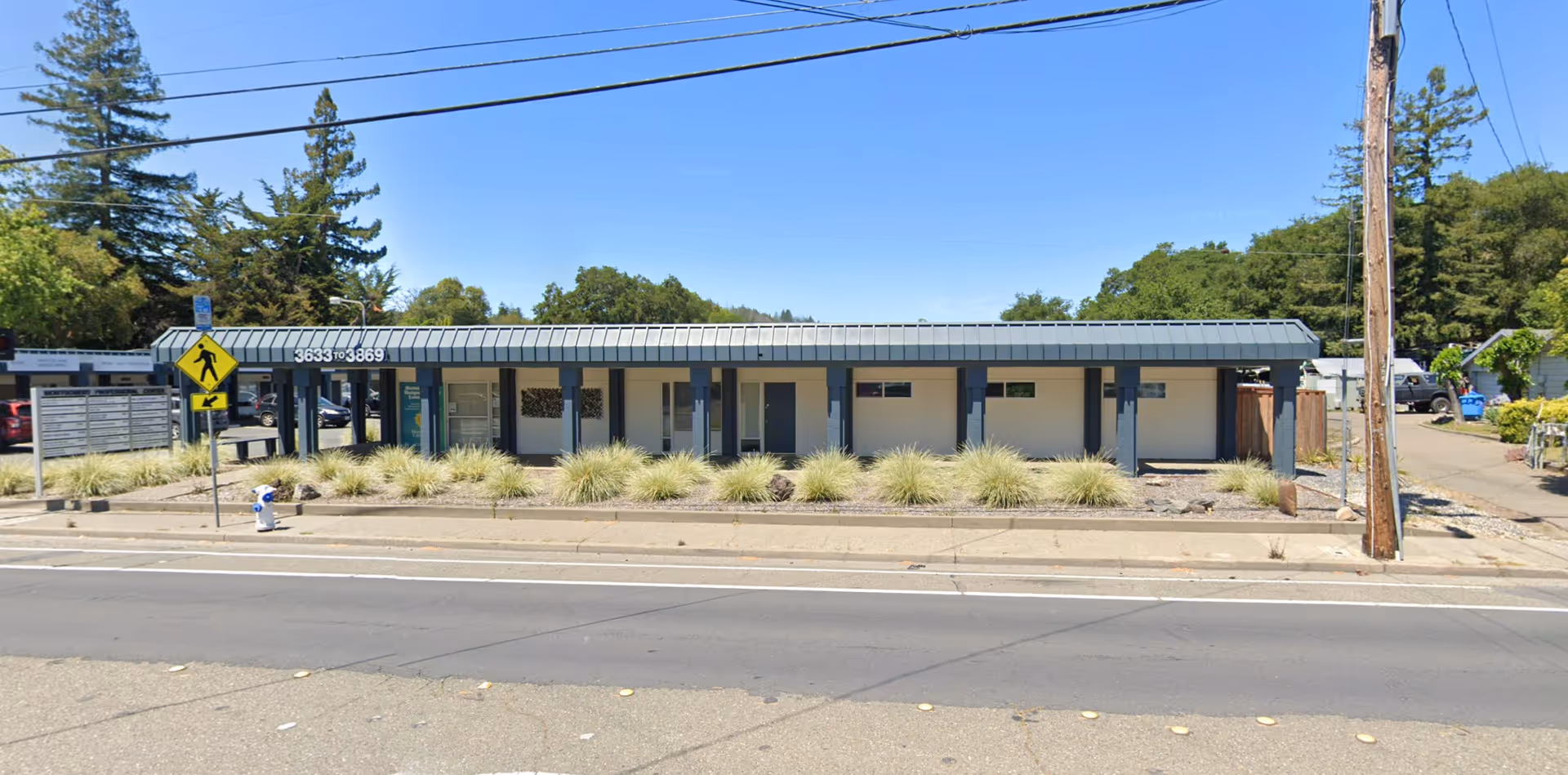 Single-story beige building with a blue canopy along the street, front landscaping with grasses.