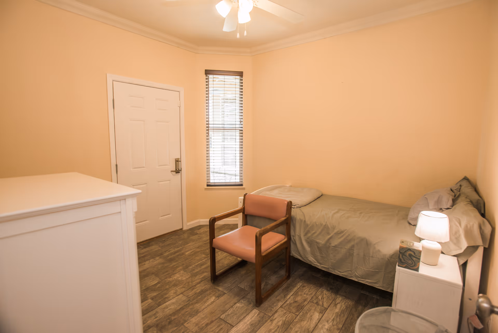A small beige bedroom with a single bed, chair, dresser, window, and bedside lamp.