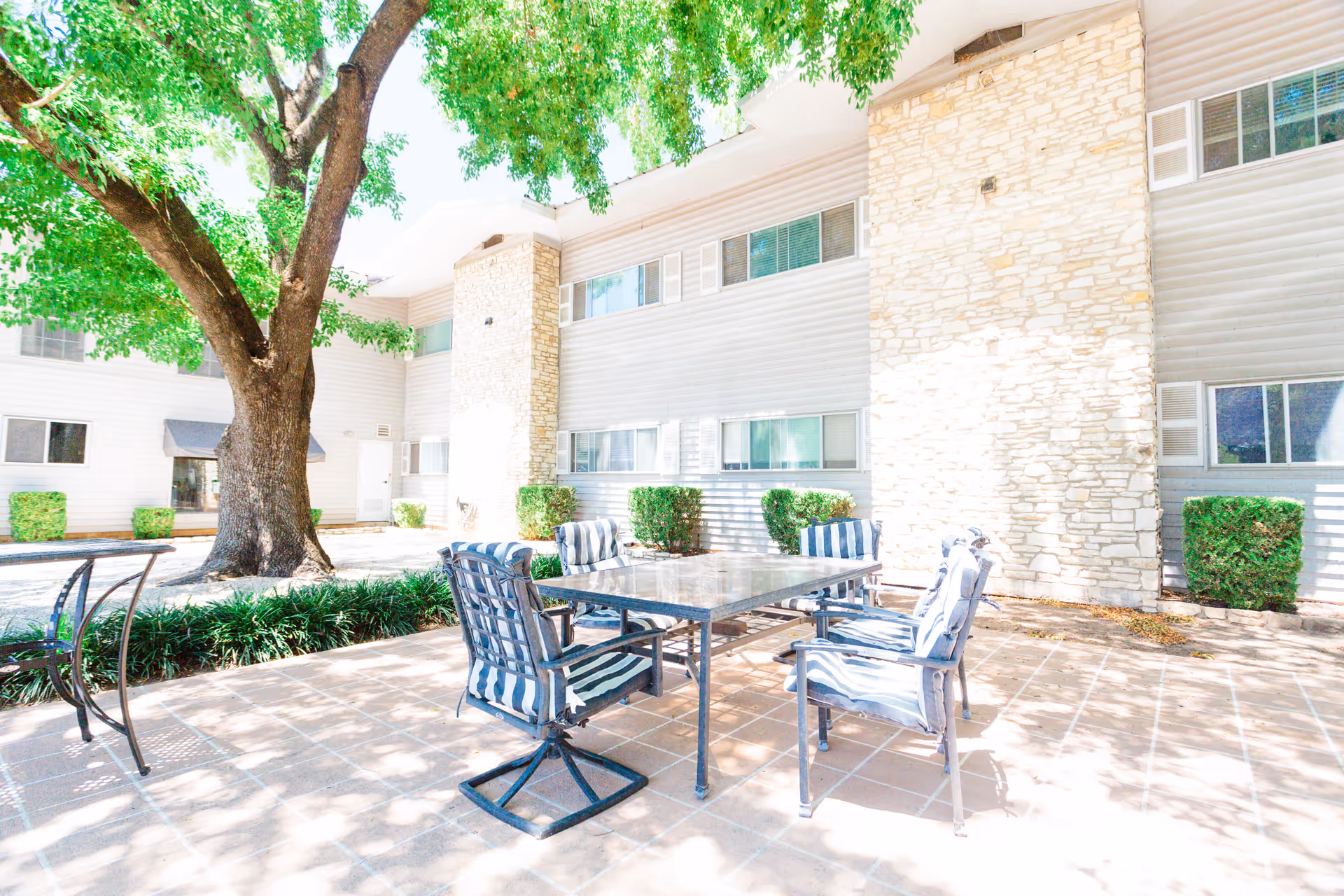 Sunny outdoor patio with a table, striped chairs, and a large tree beside a stone building.