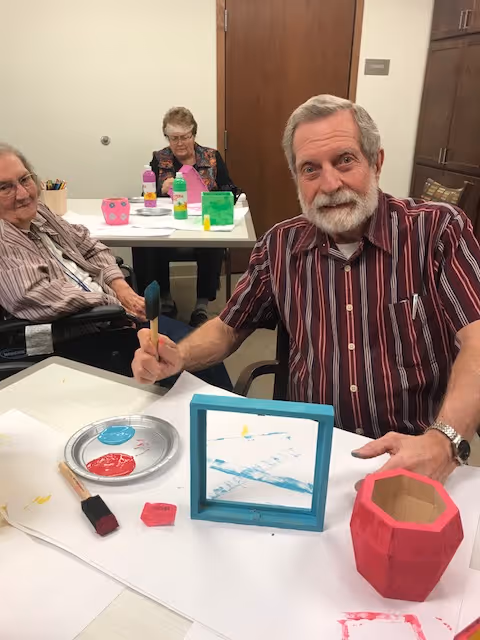 Three elderly individuals seated at tables in a room, engaging in a painting activity. One man in a striped shirt is holding a paintbrush and smiling at the camera. There are paint supplies, including a plate with red and blue paint, brushes, and painted objects on the table.
