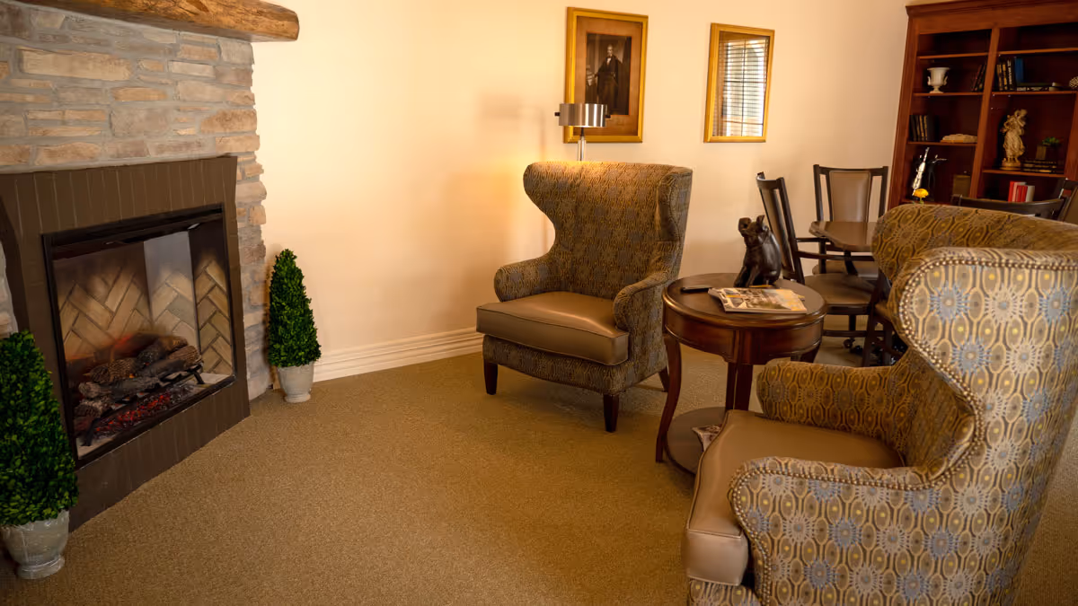A cozy living room area with two patterned wingback chairs facing a stone fireplace with a wooden mantel. Between the chairs is a round wooden side table with a small decorative sculpture and some magazines. In the background, there is a wooden dining table with chairs and a bookshelf with various items. The walls are light-colored with framed artwork hanging.