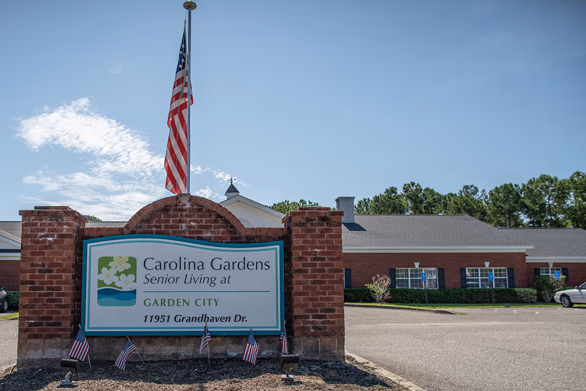 Exterior view of a senior living facility with a brick sign that reads 'Carolina Gardens Senior Living at Garden City, 11951 Grandhaven Dr.' An American flag is flying on a pole behind the sign, and there are small American flags planted in front of it. The building and parking lot are visible in the background under a clear blue sky.