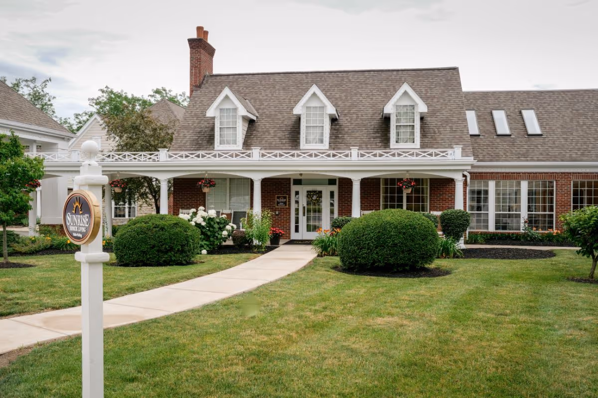 Front exterior of a brick senior living facility with a covered porch, dormer windows, landscaped lawn, and a Sunrise sign near the sidewalk.
