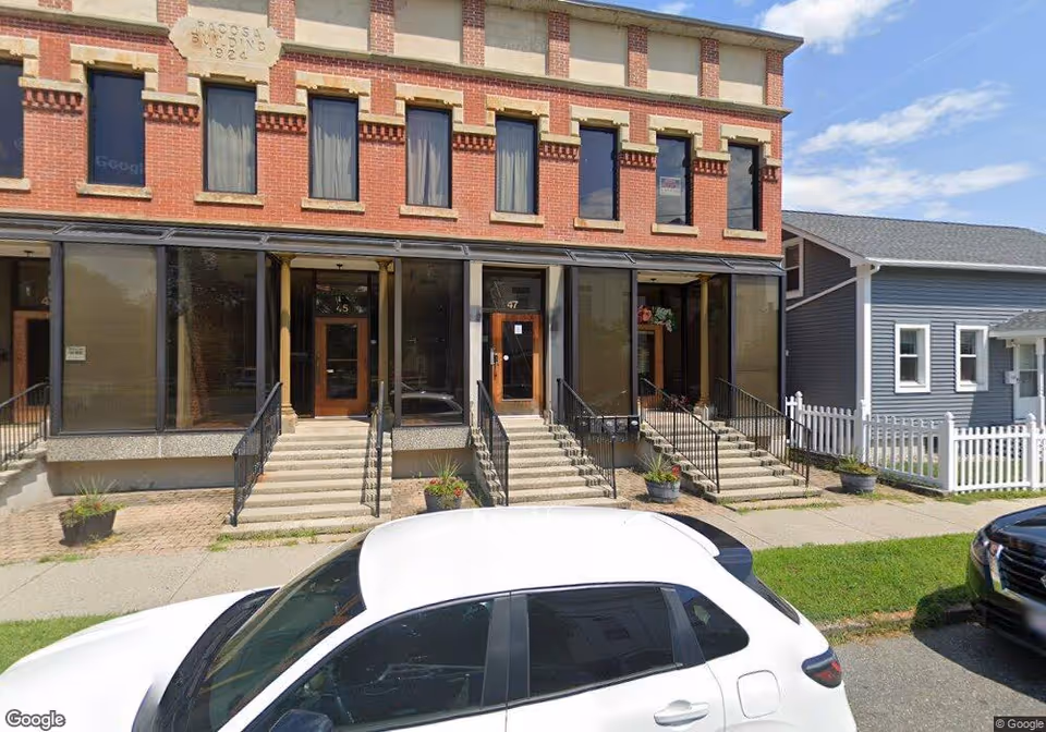 Brick storefront building with large glass windows, steps to entrances, and parked cars outside.