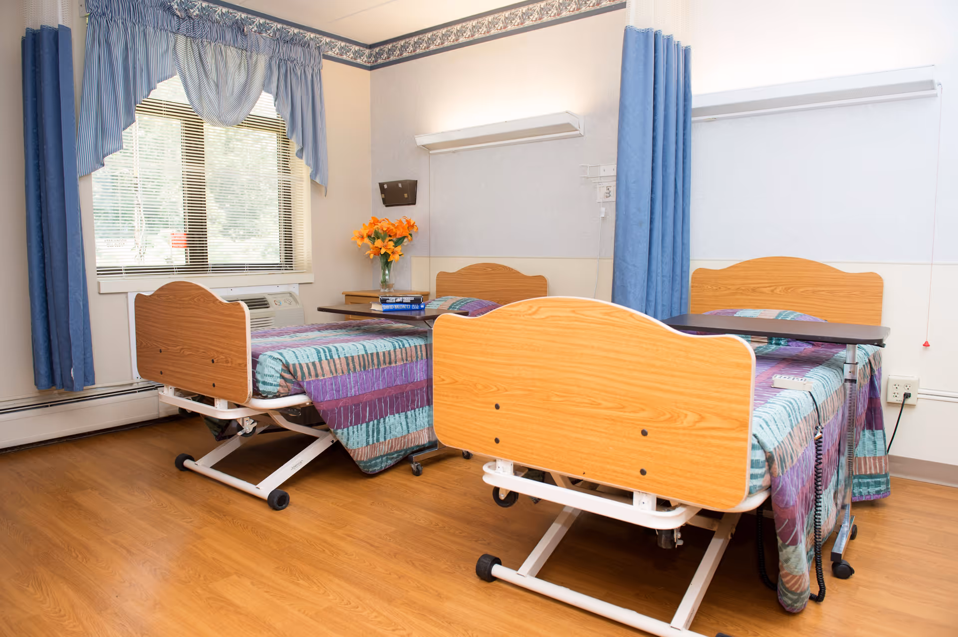 Two wooden beds in a bright shared room with blue curtains and a window.
