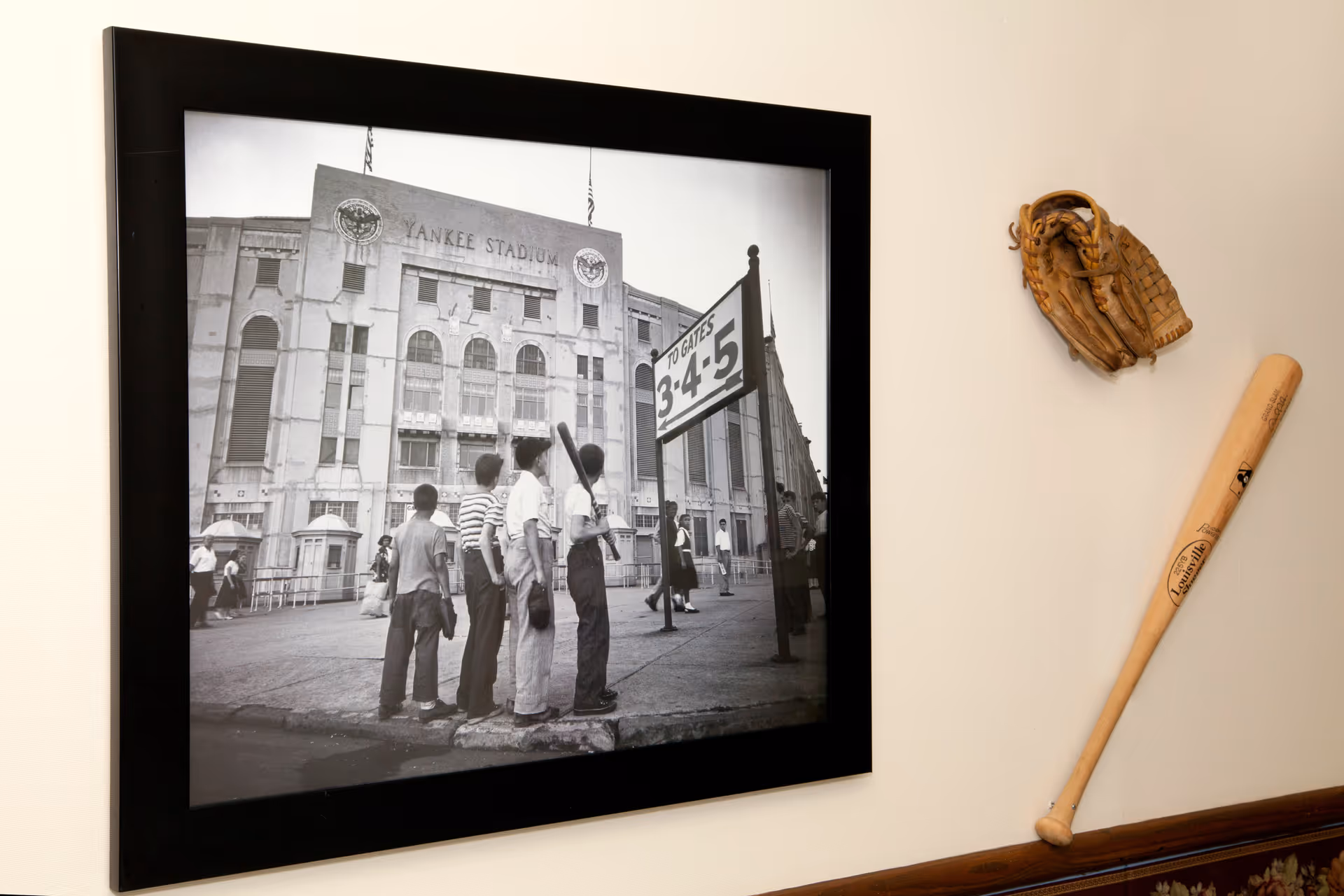 A framed black and white photograph of children standing outside Yankee Stadium, with a sign showing gates 3, 4, and 5. Next to the frame on the wall is a vintage baseball glove and a wooden baseball bat mounted as decoration.