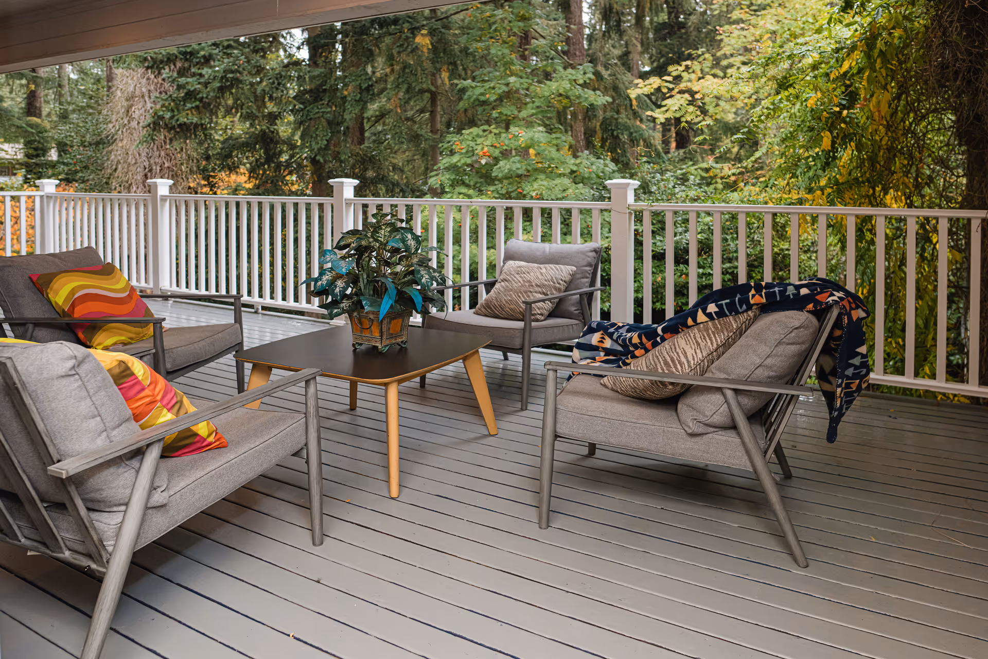 Outdoor deck seating area with chairs, a table, throw blankets, plant, and green trees.