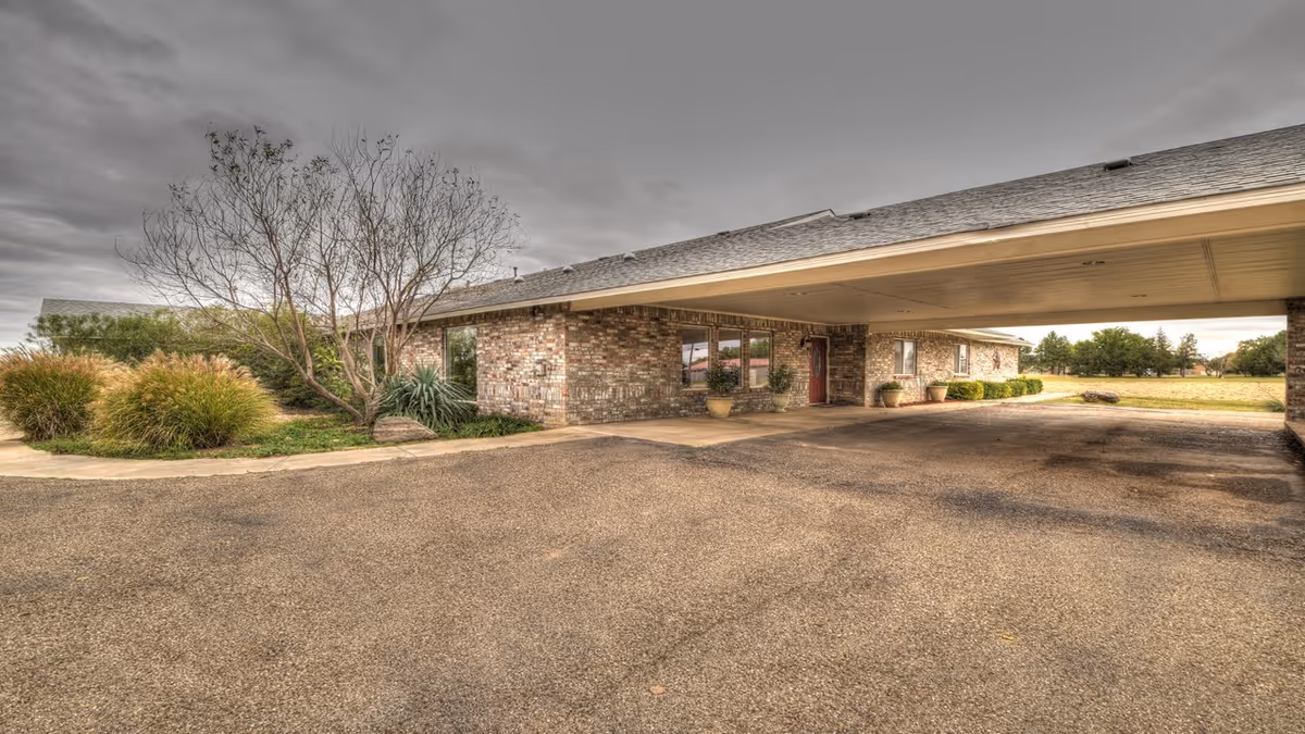 Exterior view of a single-story brick building with a covered driveway entrance, surrounded by some landscaping including bushes and a leafless tree under a cloudy sky.