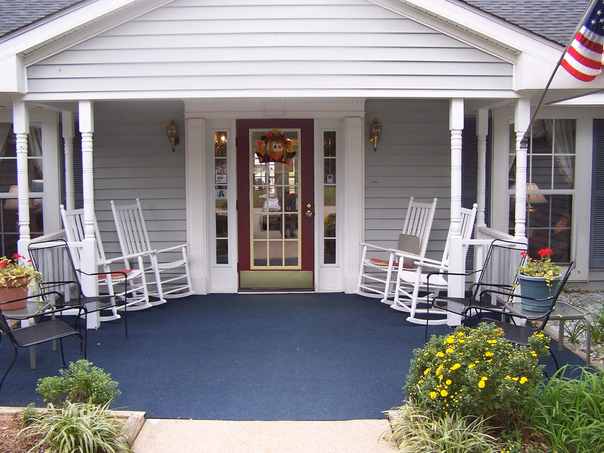 Front porch of Bayberry Retirement Inn with a maroon door featuring glass panels and a decorative wreath. The porch has white rocking chairs and black metal chairs with potted plants and flowers on either side. An American flag is visible on the right side near a window with blue shutters.
