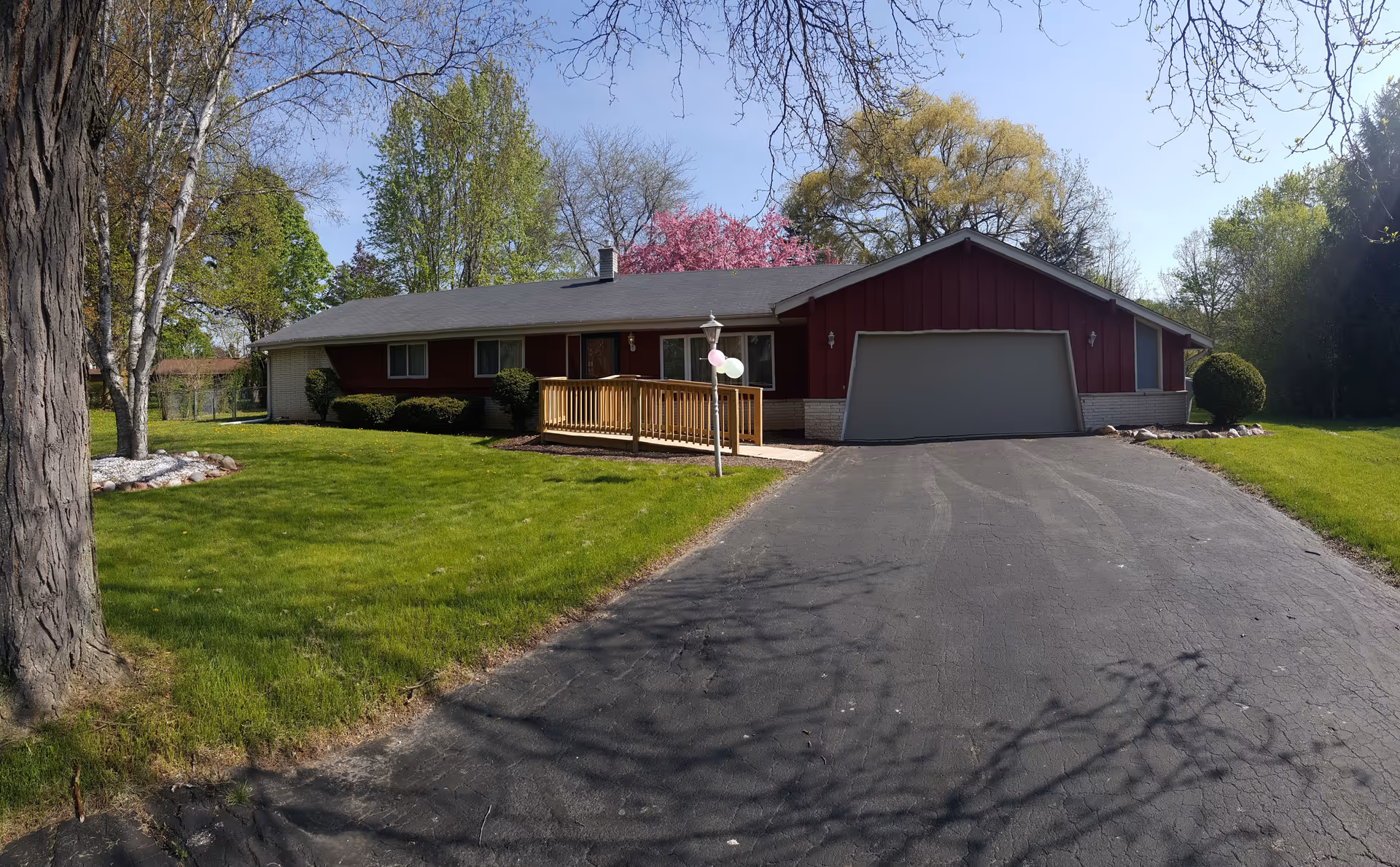 Single-story residential building with a red and white exterior, a two-car garage, a wooden ramp leading to the front door, and a well-maintained lawn with trees and bushes under a clear blue sky.