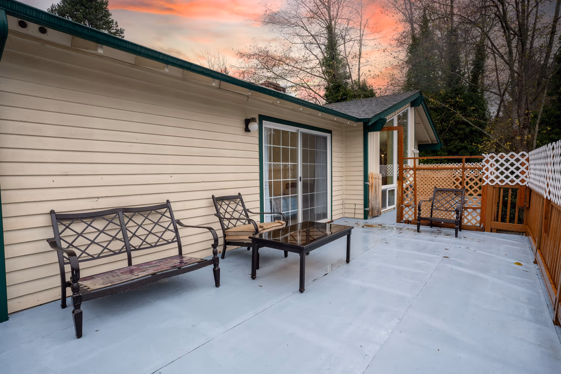 Outdoor patio attached to a beige house with benches, a table, and lattice fencing.