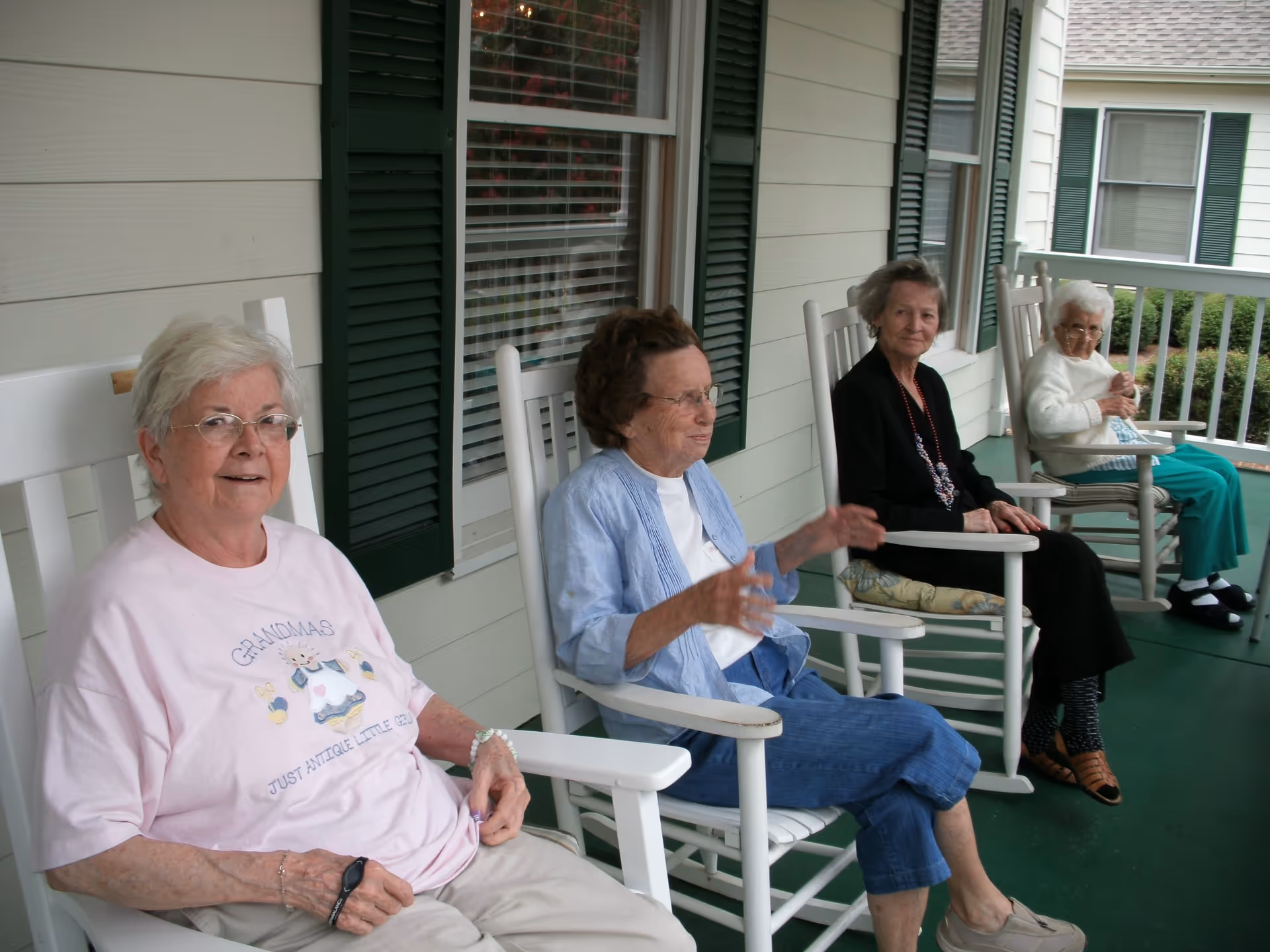Four elderly women sitting on white rocking chairs on a covered porch with green floor and white railing. The porch is attached to a building with beige siding and green shutters on the windows.