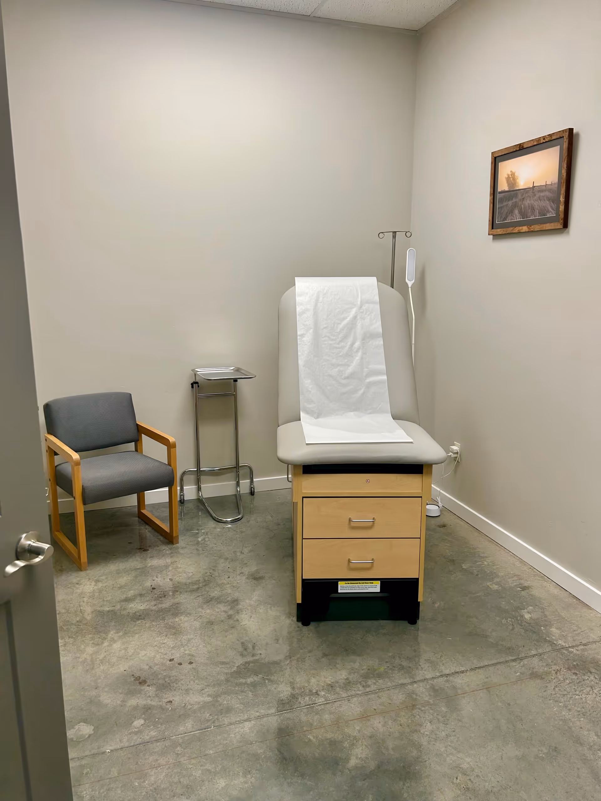 Examination room with a medical chair, paper sheet, cabinet, IV stand, small chair, framed wall photo.