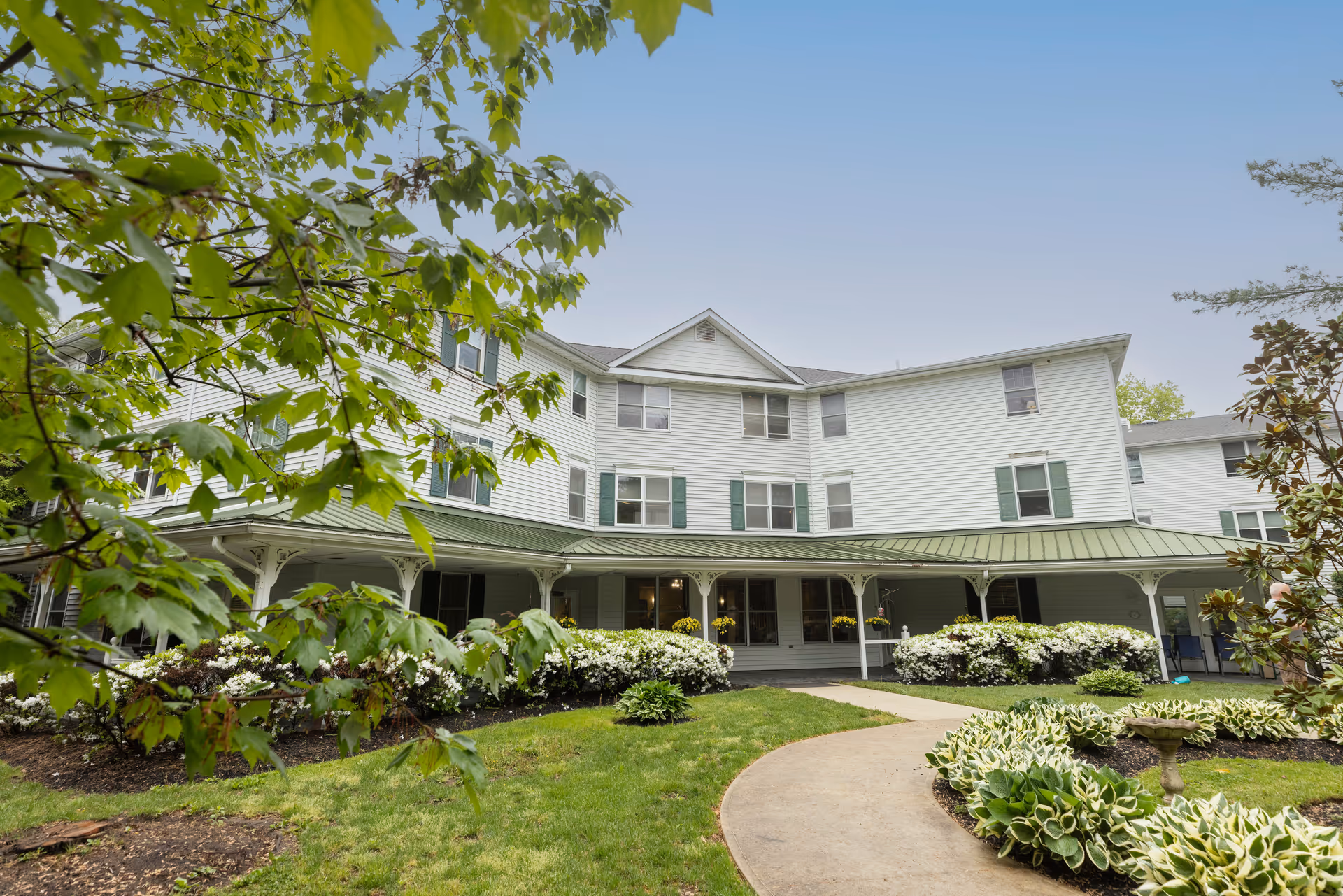 Front exterior of a white multi-story senior living building with a covered porch, landscaped gardens, and a curved walkway.