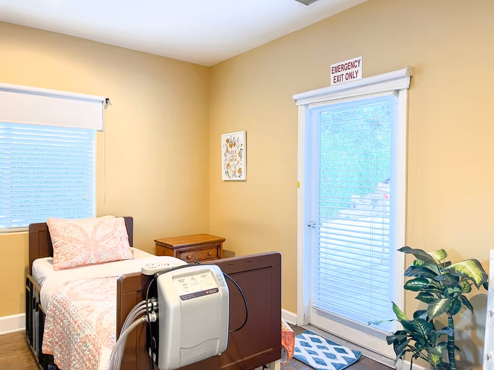 Single-occupancy bedroom with a bed, a medical device at the foot of the bed, a wooden nightstand, a potted plant, window blinds, and a door labeled "EMERGENCY EXIT ONLY".