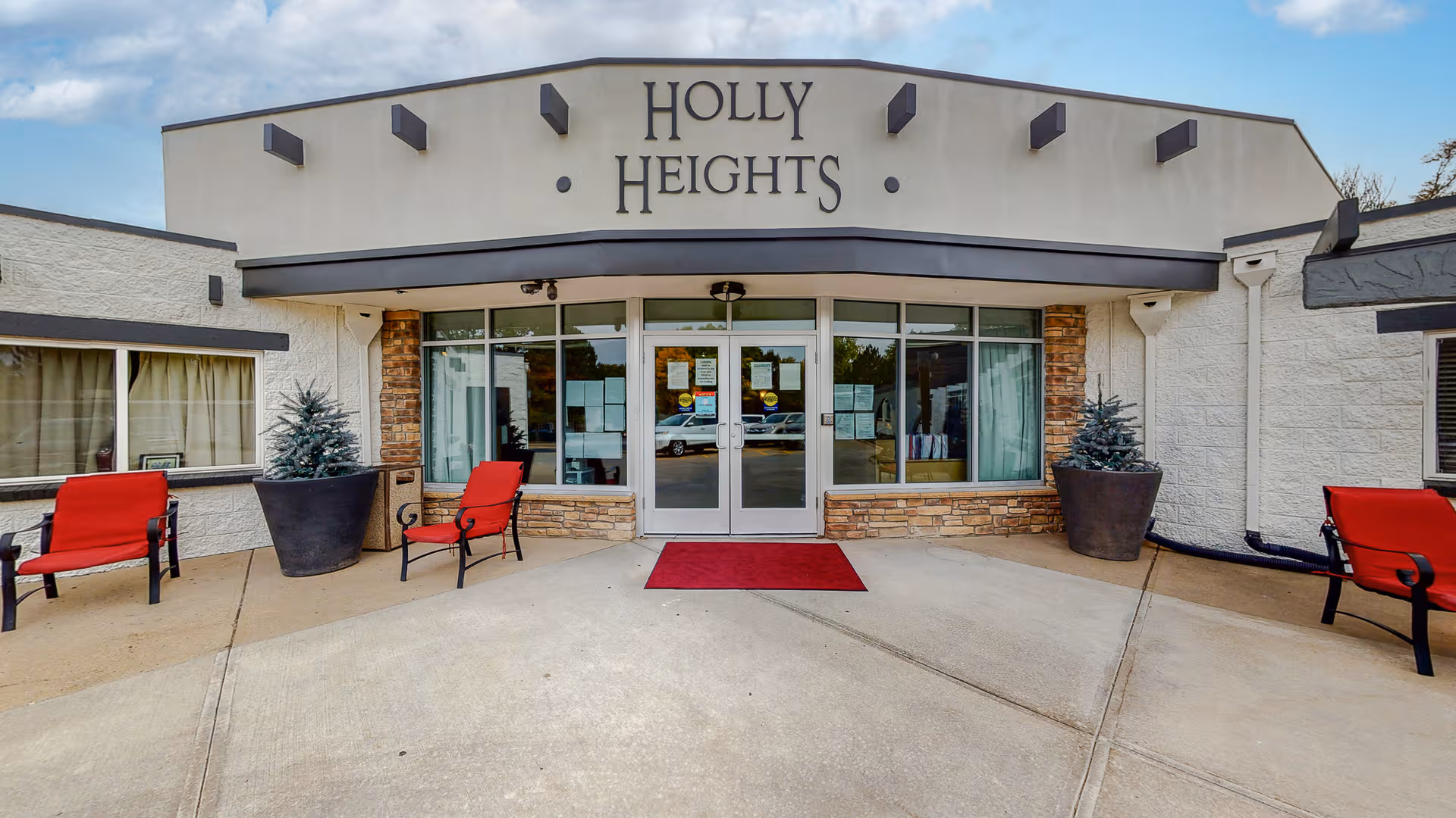 Front entrance of Holly Heights with glass doors, red chairs, and large potted plants.