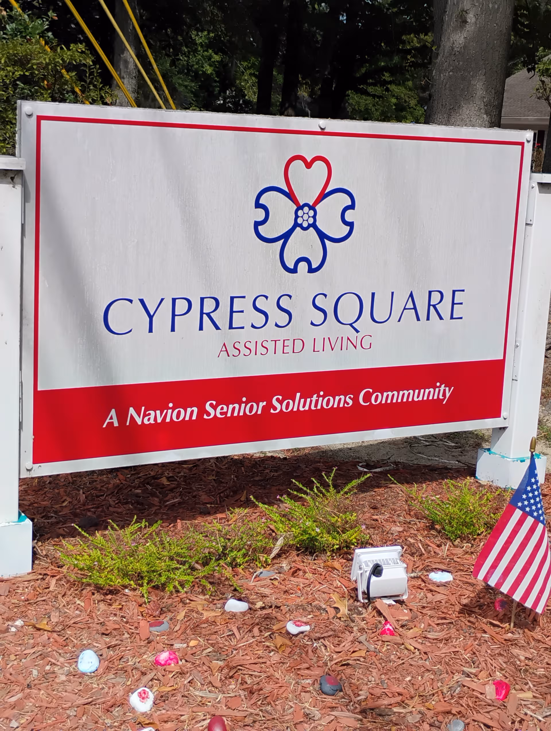 Exterior sign reading Cypress Square Assisted Living with a flower logo and an American flag nearby.