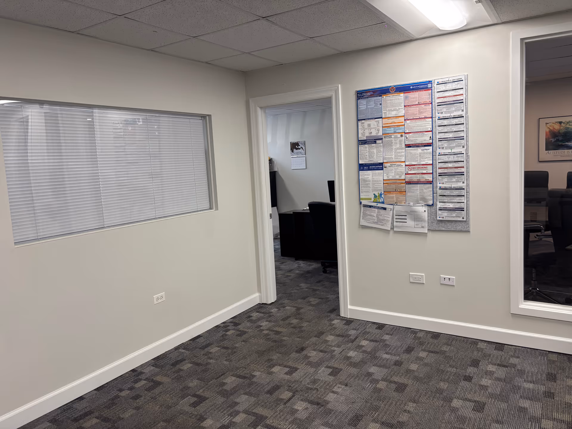 Interior facility hallway with windowed wall, doorway to a meeting room, and a large bulletin board.