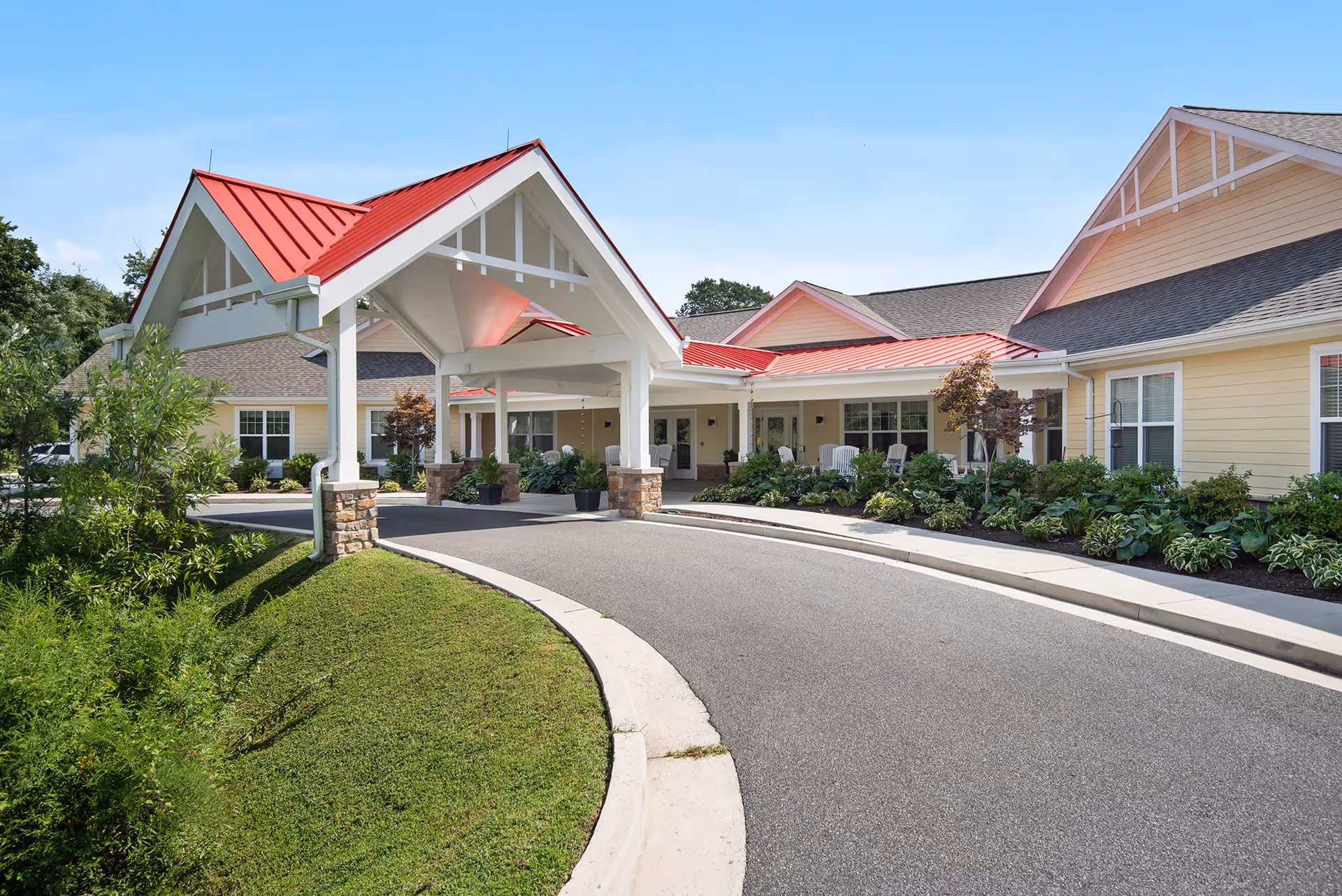 Exterior view of a senior living facility named Candle Light Cove featuring a covered entrance with a red roof, a curved driveway, well-maintained landscaping with bushes and small trees, and a clear blue sky.