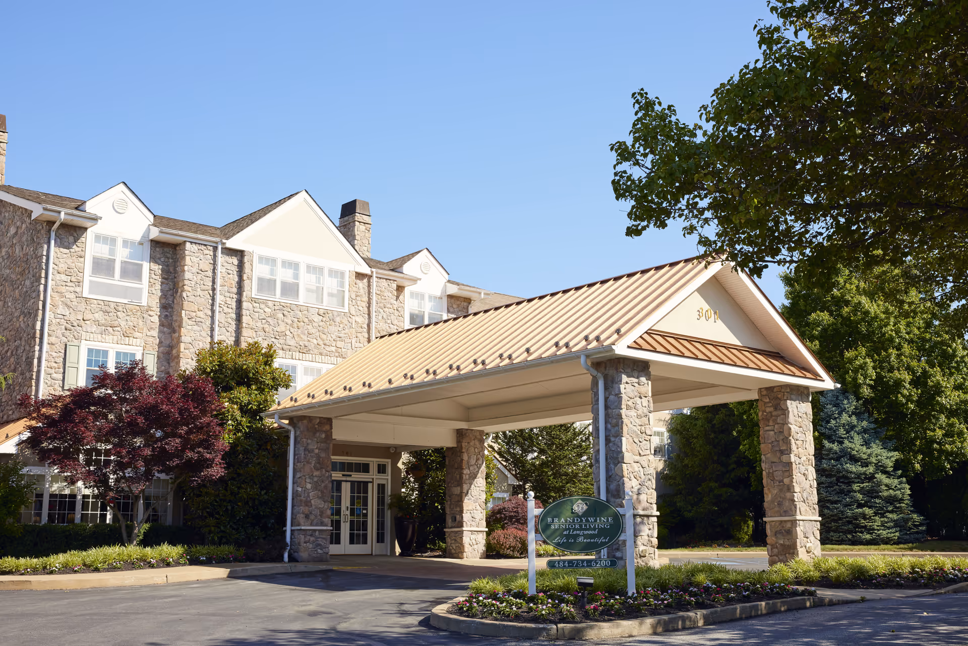 Exterior view of Brandywine Longwood by Monarch senior living facility showing the entrance with a covered driveway supported by stone pillars, surrounded by trees and landscaping under a clear blue sky.