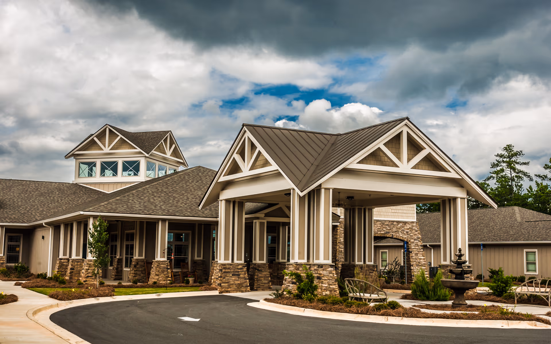 Exterior view of Fellowship Meriwether Assisted Living Community showing a covered entrance with stone pillars, a circular driveway, landscaped greenery, and a multi-gabled roof under a cloudy sky.