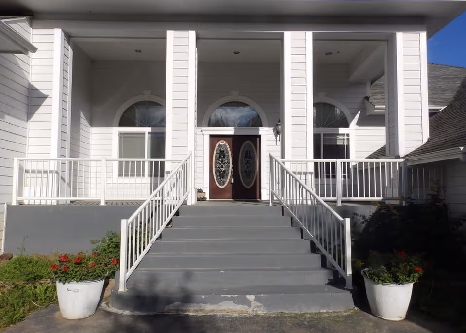 Front entrance of a white building with double doors, stairs, railings, and potted plants.