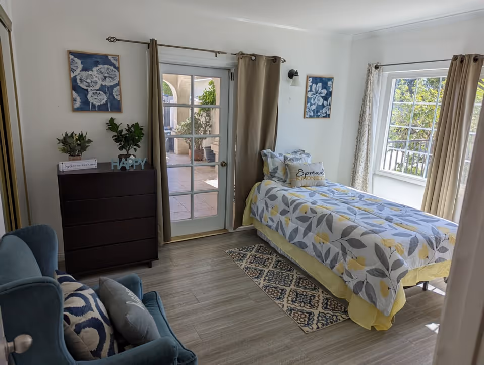 Bright bedroom with patterned bedspread, dresser, curtains, and a blue chair.