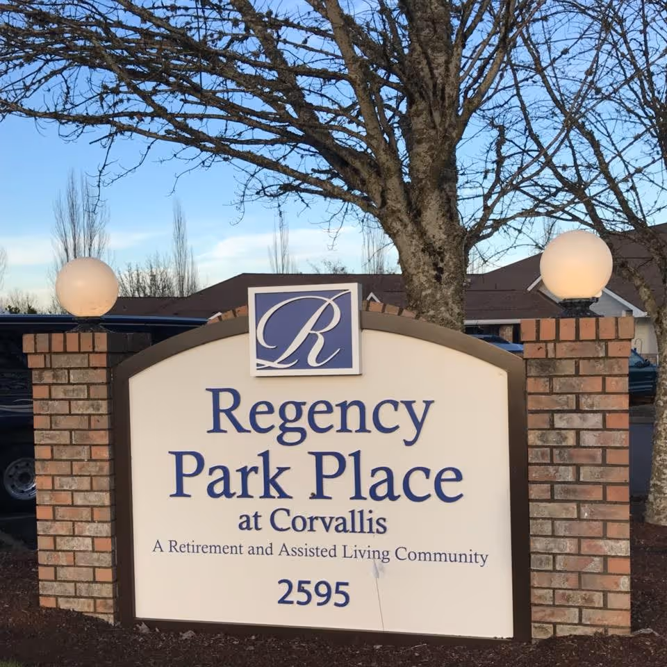 Brick entrance sign reading "Regency Park Place at Corvallis, A Retirement and Assisted Living Community, 2595" with trees and globe lights behind it.