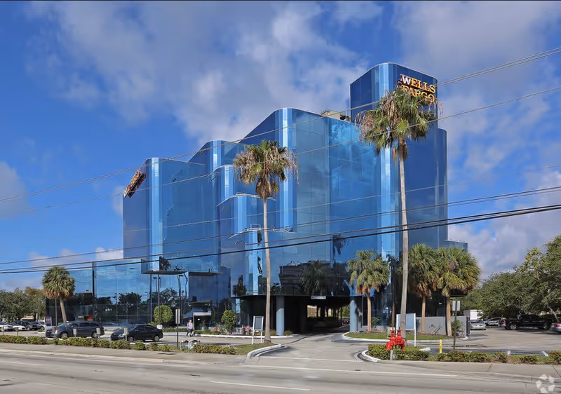 Blue glass multi-story building with palm trees along a street under a sunny sky.