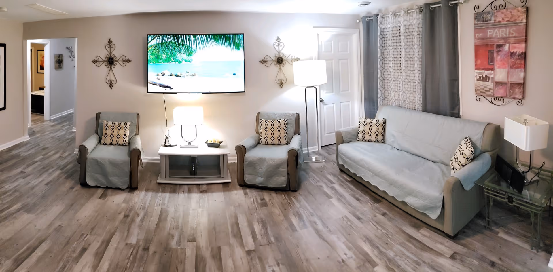 A cozy living room with two armchairs and a sofa covered with light blue slipcovers, each with patterned throw pillows. A flat-screen TV is mounted on the wall above a small white table with a lamp and decorative items. The room has wood-look flooring, neutral-colored walls, decorative wall art including two metal crosses and a Paris-themed picture, and a floor lamp near a closed white door. Sheer and solid curtains cover the window on the right side.