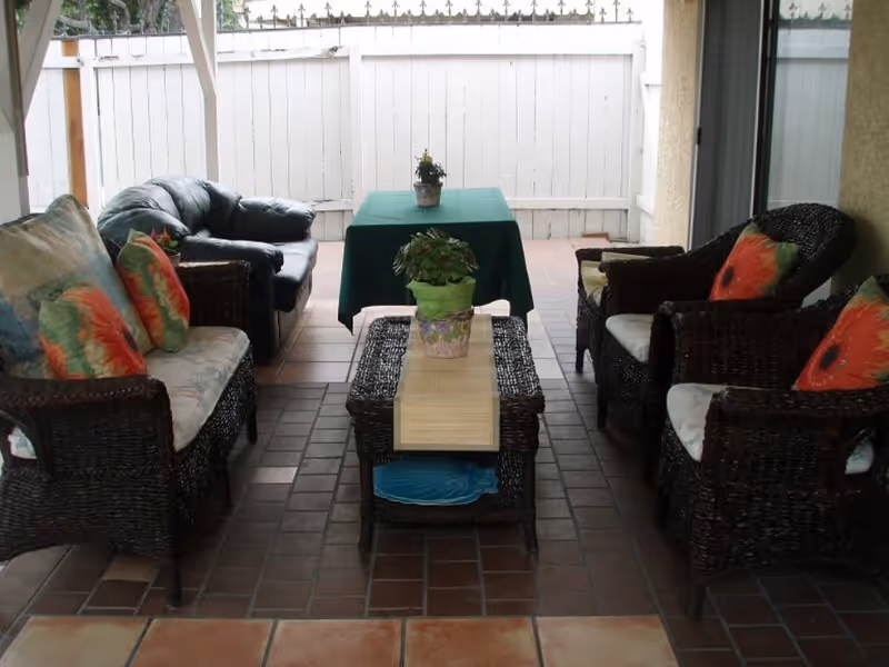 Covered outdoor patio area with wicker furniture including a loveseat, two armchairs with floral cushions, and a black leather sofa. A rectangular coffee table with a potted plant sits in the center, and a table covered with a green cloth and another potted plant is against a white wooden fence in the background.