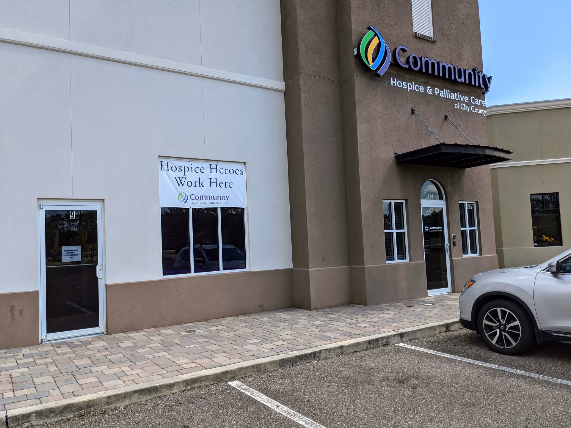 Exterior of a beige building with Community Hospice signage, banner window, and parked car.