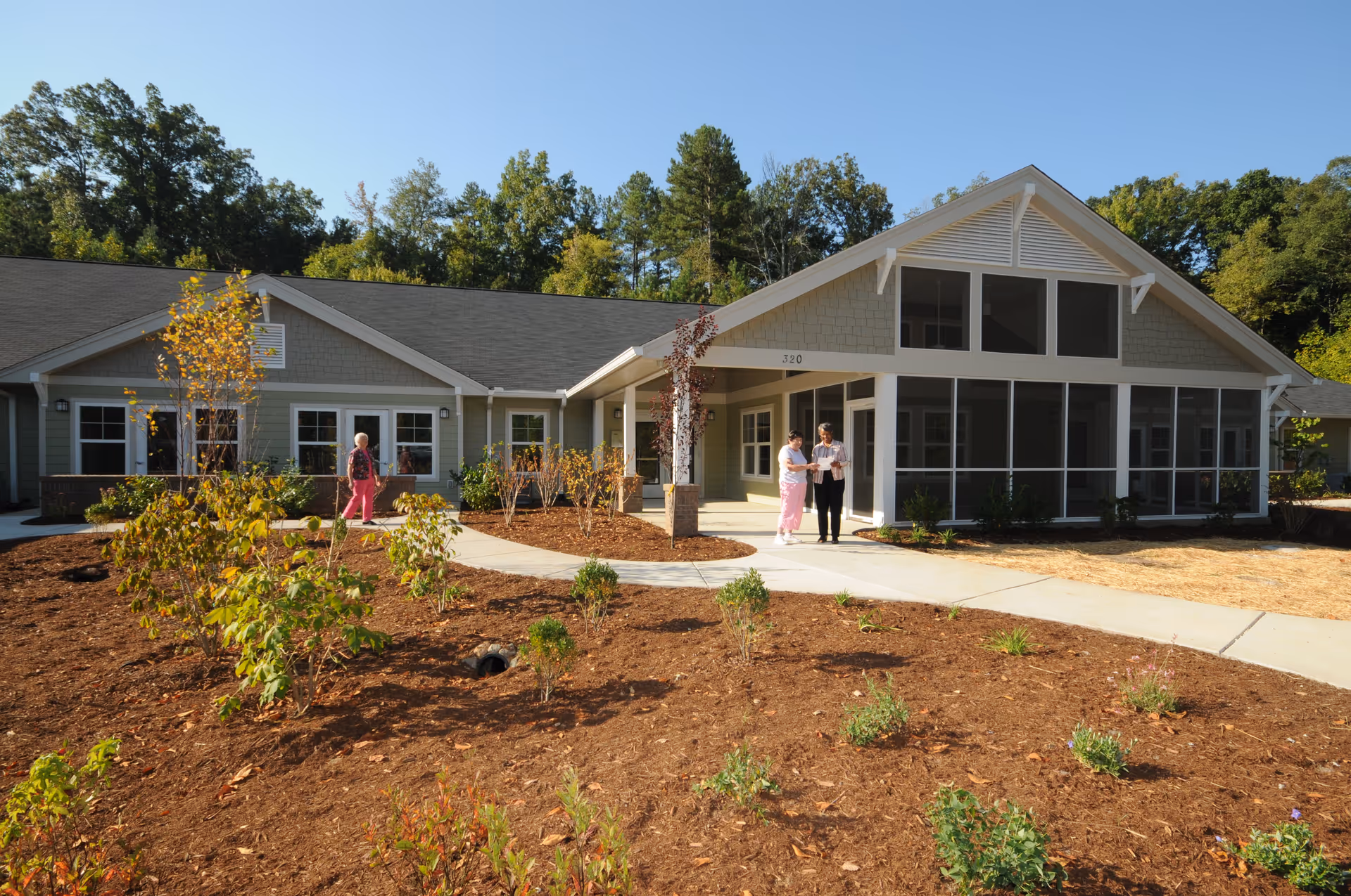 Exterior view of a single-story senior living facility building with a sloped roof and large windows. There are two people standing and talking near the entrance, and another person walking on the pathway. The landscaping includes small plants and trees with mulch covering the ground. The sky is clear and blue.