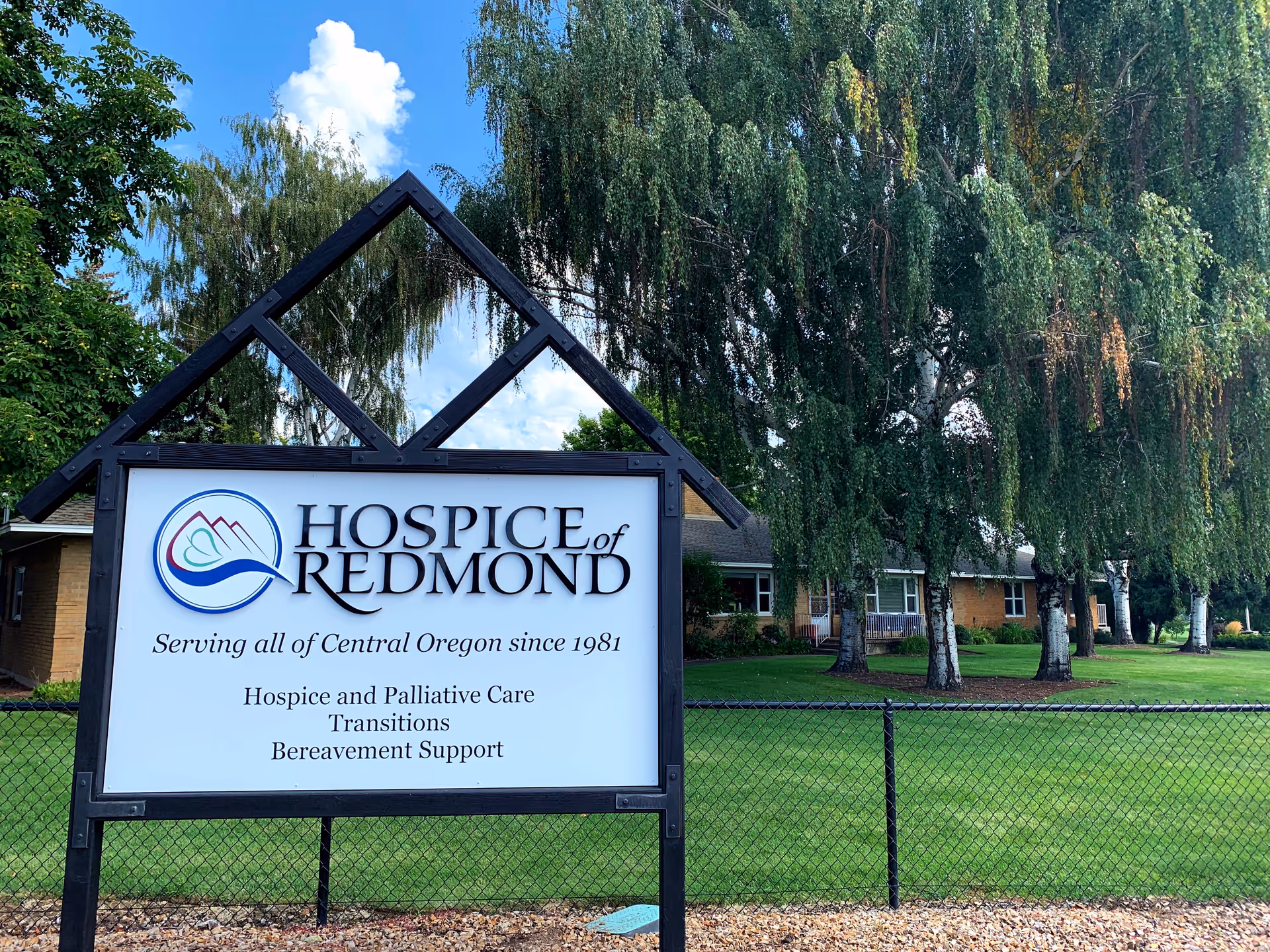 Front exterior sign for Hospice of Redmond with lawn, trees, and a fence.