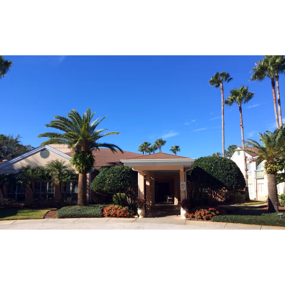 Front entrance of a single-story senior living building with a covered porte-cochere, palm trees and manicured landscaping under a clear blue sky.