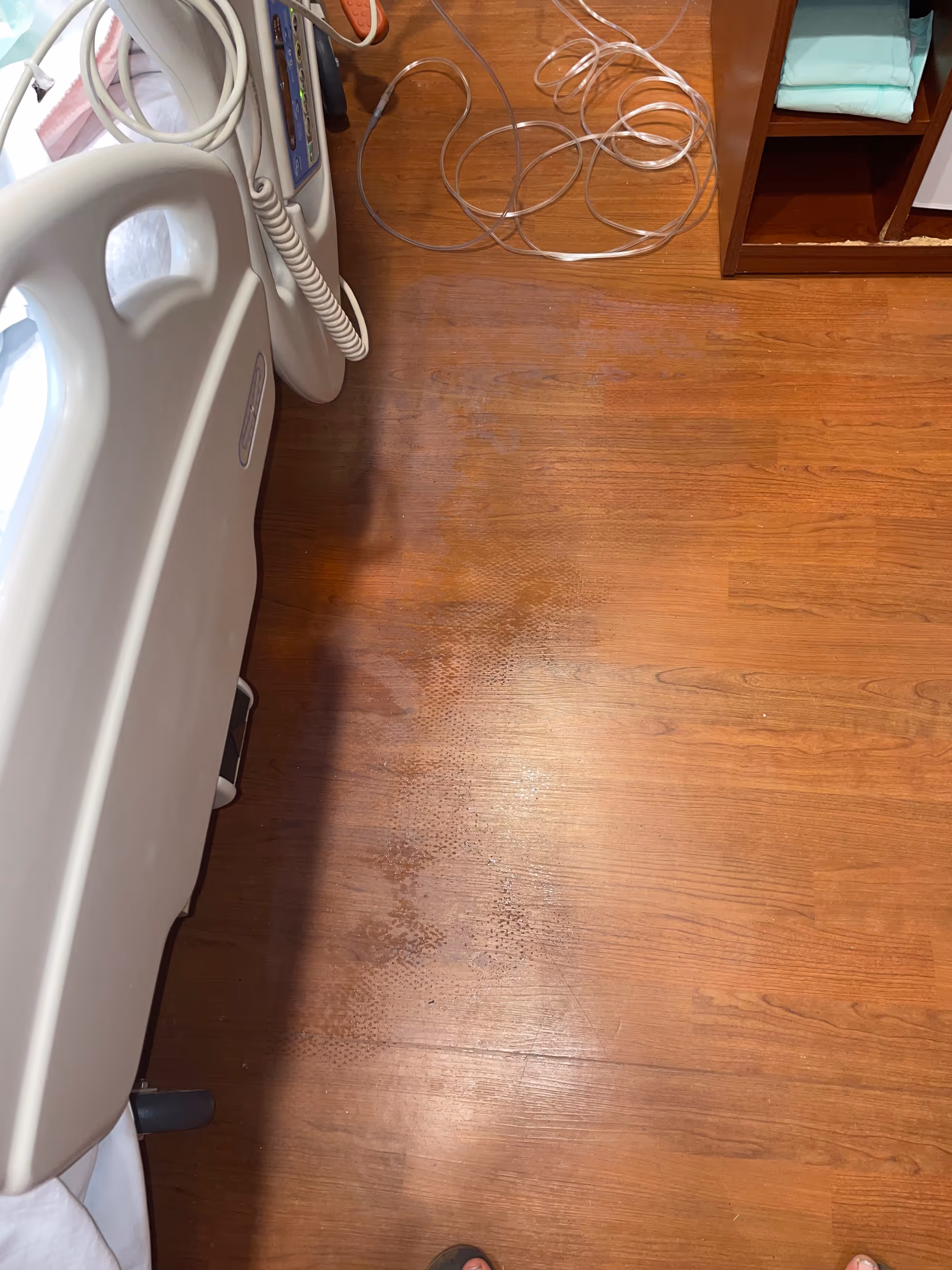 Hospital bed with tangled cords on a wooden floor beside a shelf in a patient room.