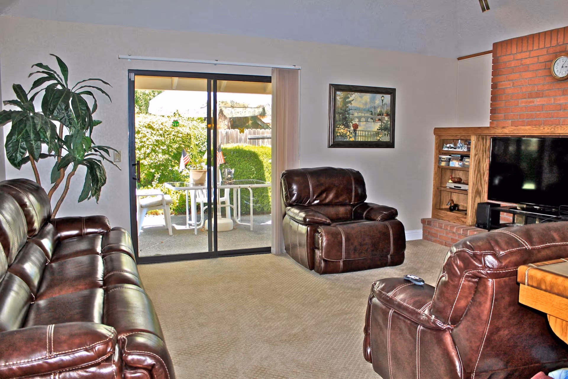 Living room with brown leather sofa and armchairs, a television and brick fireplace, and a sliding glass door opening to a patio.