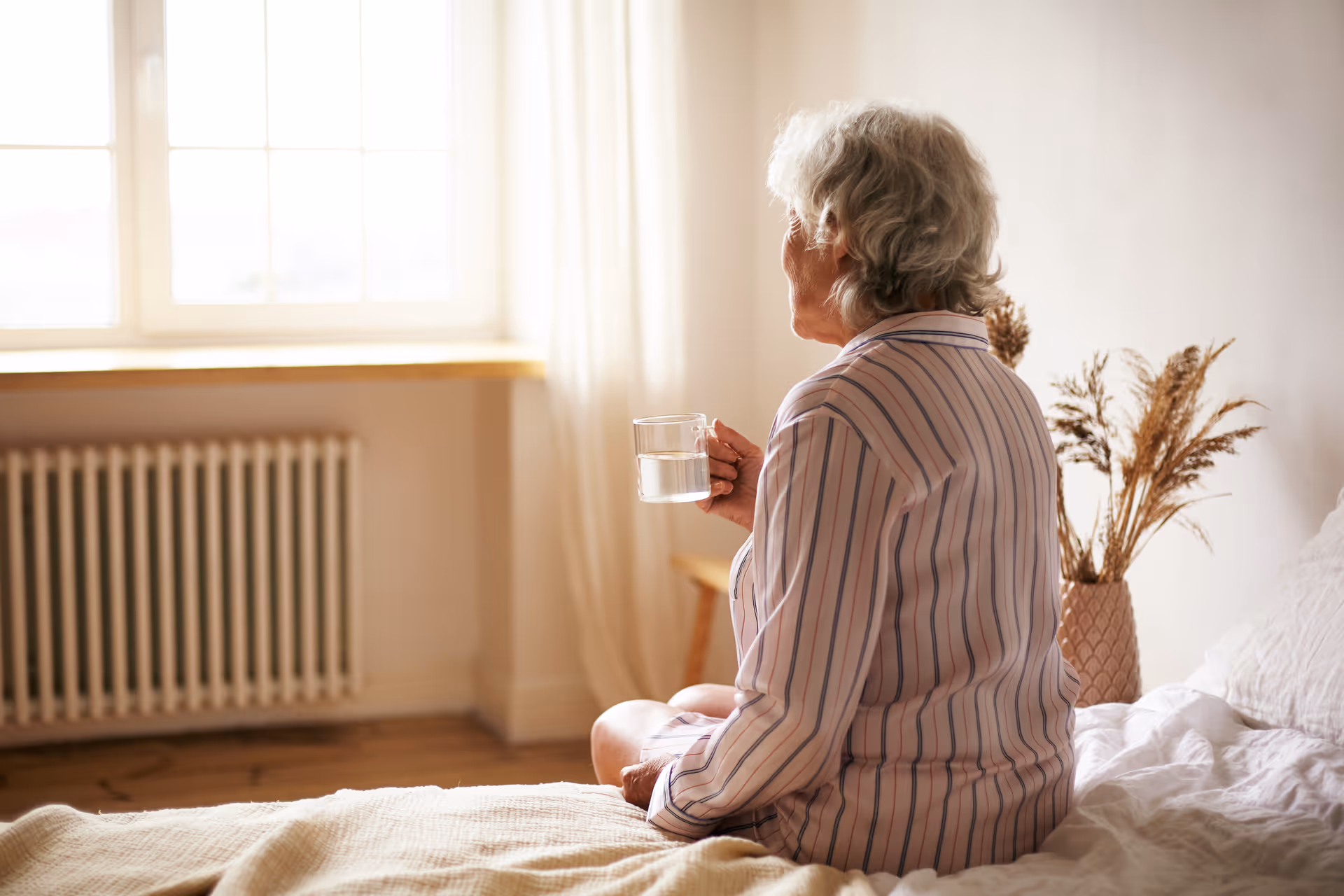 Elderly woman sits on a bed in a sunlit bedroom, holding a glass of water.