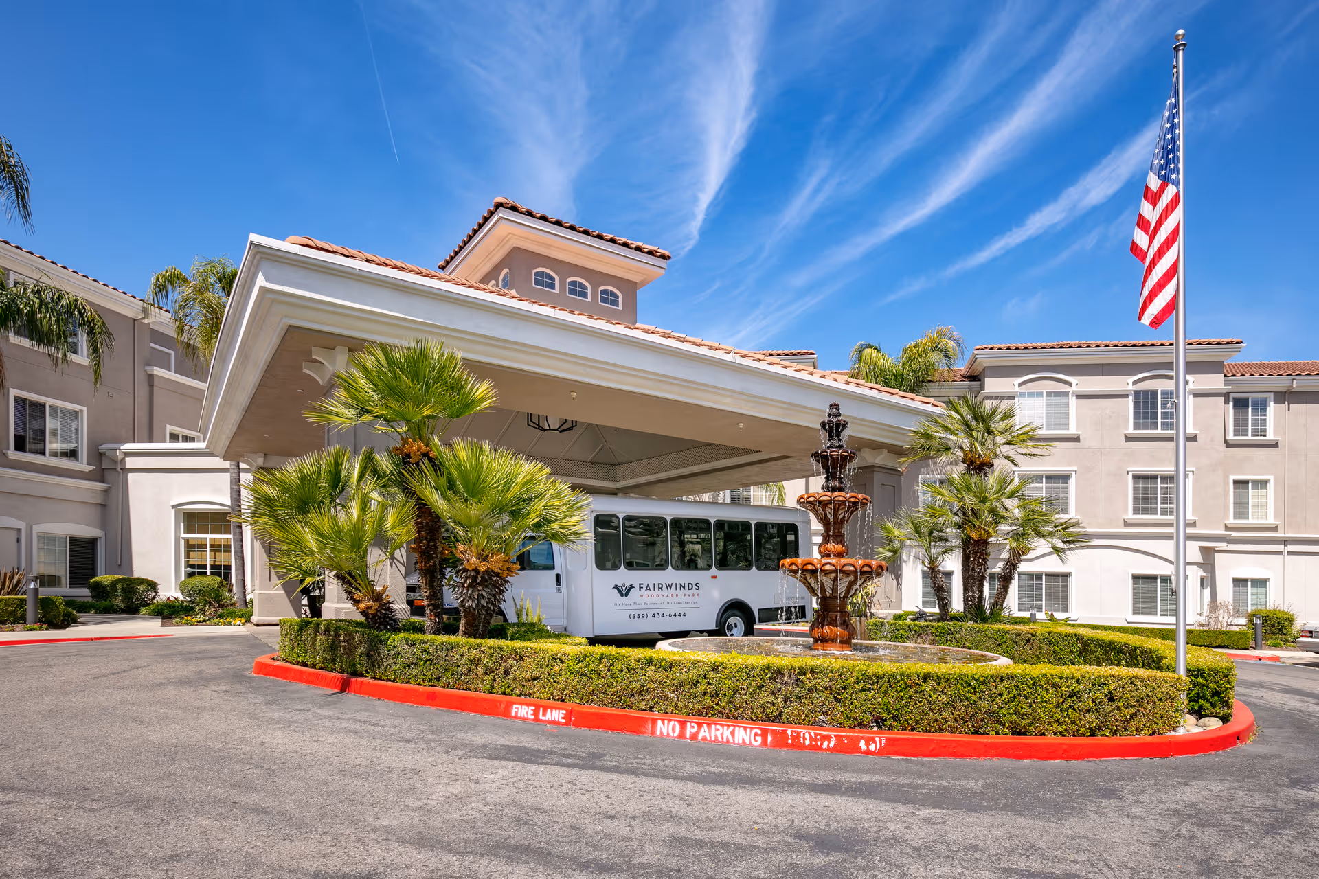 Exterior view of Fairwinds Woodward Park with front entrance canopy, palm trees, fountain, and bus.